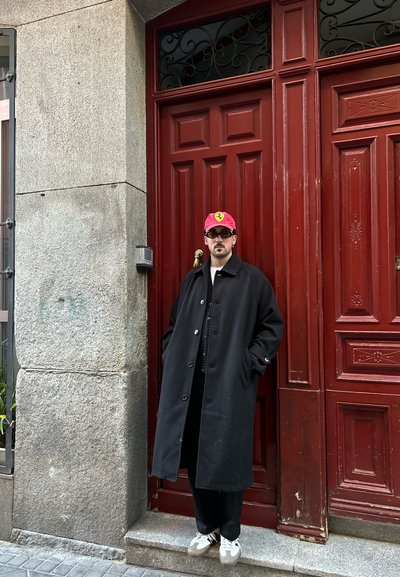 Abrigo negro con silueta oversized, usado sobre un atuendo oscuro. Gorra de béisbol roja con logo, gafas de sol oscuras y zapatillas ligeras. Fondo de puerta de madera roja.