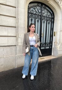Överdimensionerad rutiga kavaj, vit linne, ljusblå vida jeans, grå och vita sneakers, liten krämfärgad crossbody-väska, stående nära en utsmyckad dörr.