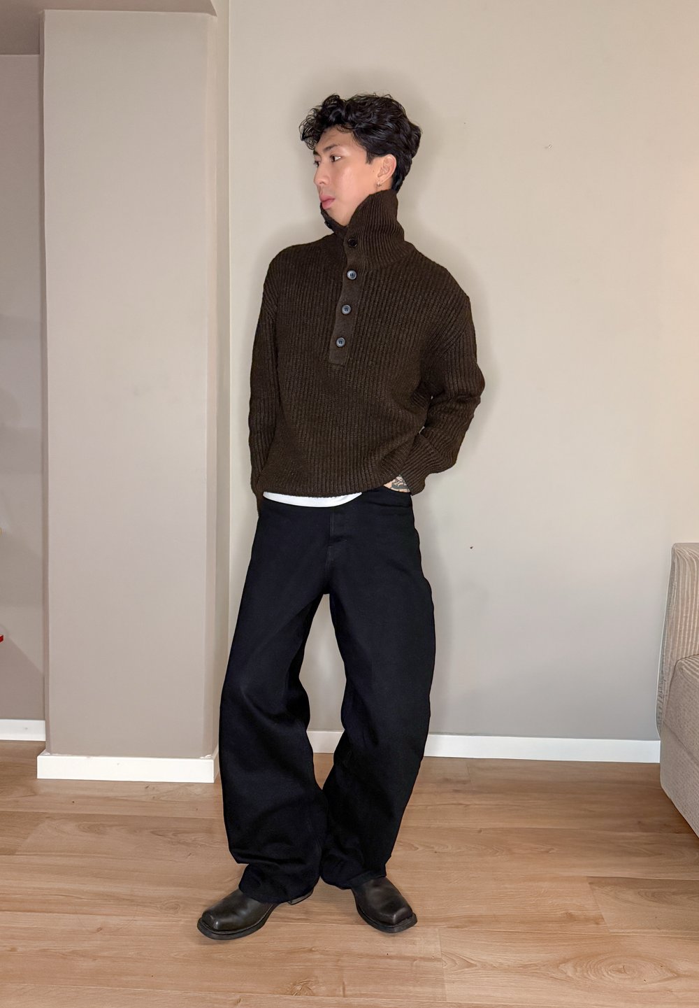Pull côtelé brun foncé à col montant avec trois boutons, associé à un pantalon noir ample et des chaussures noires, sur un fond de mur neutre.