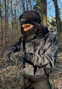 Giacca nera imbottita con cappuccio foderato di pelliccia, scaldacollo a strati e guanti. Tessuto lavorato e polsini elasticizzati. Sfondo di foresta, ambientazione invernale.
