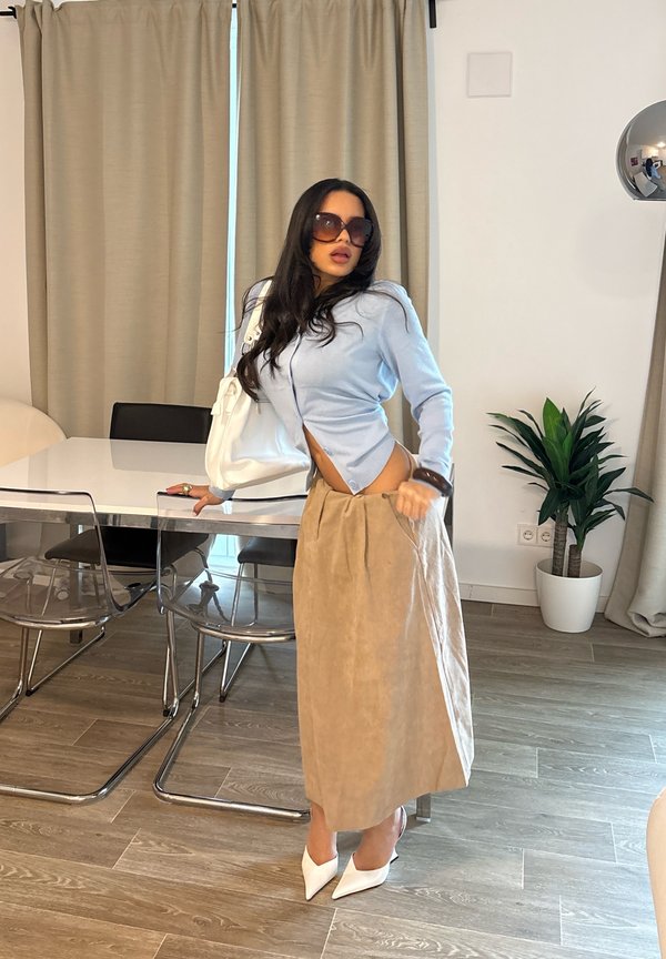 Vrouw met lang donker haar, zonnebril, lichtblauw topje, beige rok en witte hakken, staande naast een eettafel in een moderne kamer.