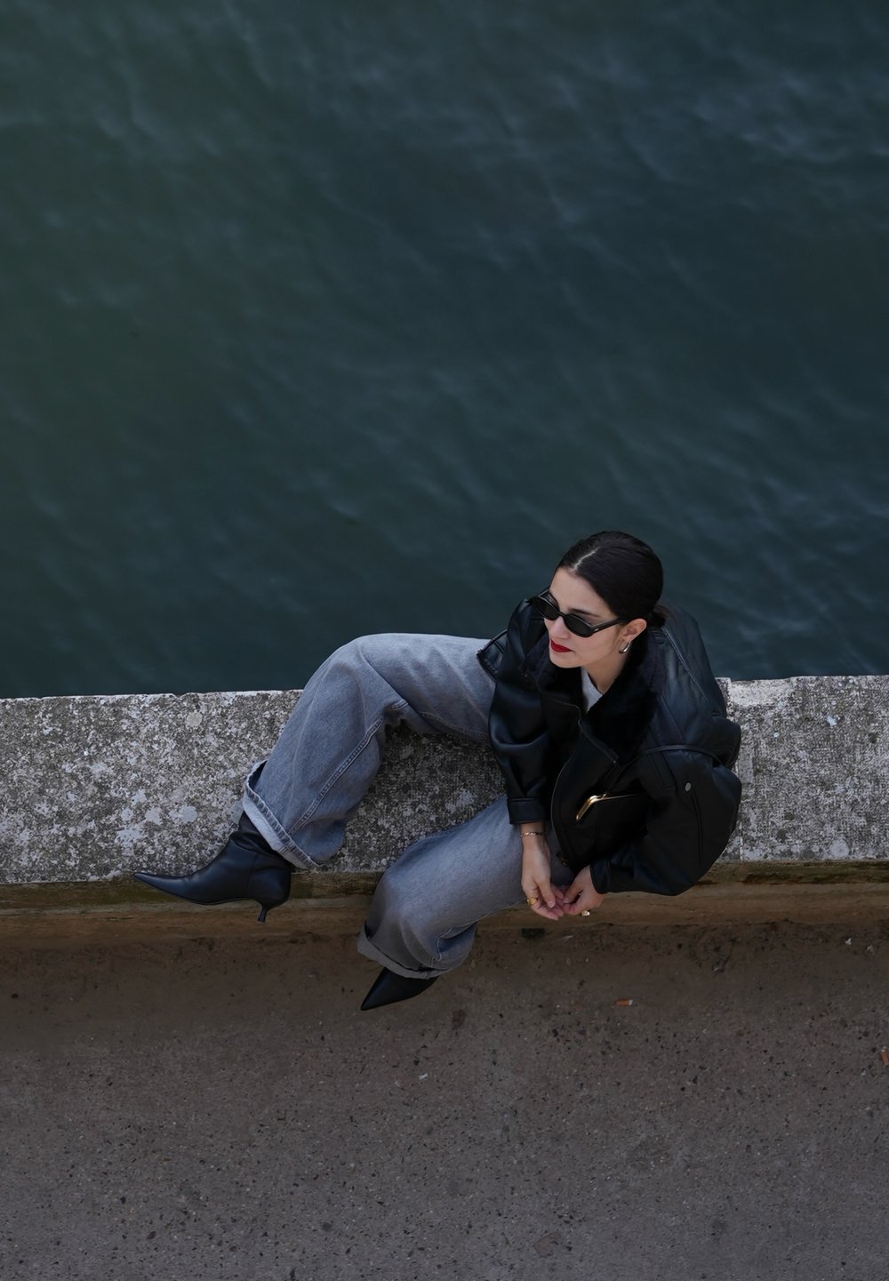 Schwarze Lederjacke, graue übergroße Jeans und schwarze High-Heels. Accessoires umfassen Sonnenbrillen und eine schwarze Tasche. Wasserkulisse.