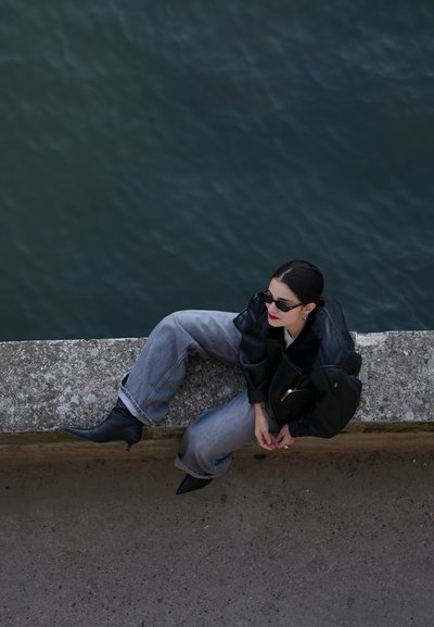 Chaqueta de cuero negra, jeans gris oversized y botas negras de tacón alto. Los accesorios incluyen gafas de sol y un bolso negro. Fondo acuático.