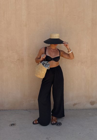 Top de bikini negro y pantalones negros de pierna ancha. Sombrero de paja de ala ancha y un bolso de mimbre. Joyas simples como acentos. Fondo neutro.