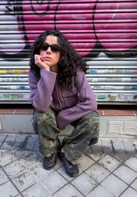 Femme aux cheveux bouclés foncés et lunettes de soleil, accroupie sur un trottoir carrelé, portant un pull violet et un pantalon camouflage devant un volet métallique couvert de graffitis.