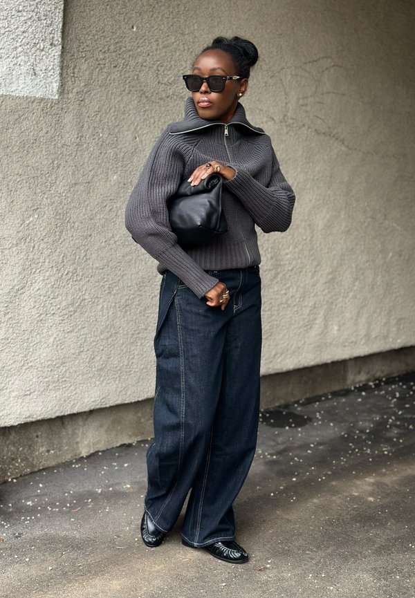 Donna con occhiali da sole scuri, maglione grigio con zip, jeans a gamba larga, scarpe nere e una pochette in pelle nera, in piedi contro un muro testurizzato.