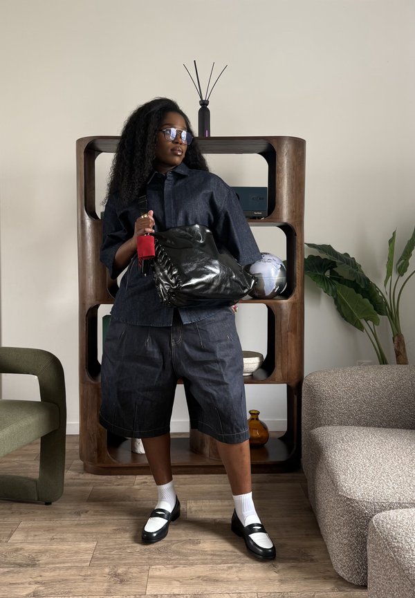 Kvinde i mørkt denimoutfit og sort-hvide sko står med en stor sort taske i en moderne stue med en træhylde og planter.