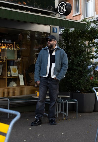 Hombre con una chaqueta de mezclilla azul sobre un suéter blanco y negro, jeans oscuros y zapatos negros, de pie frente a una tienda con vegetación.