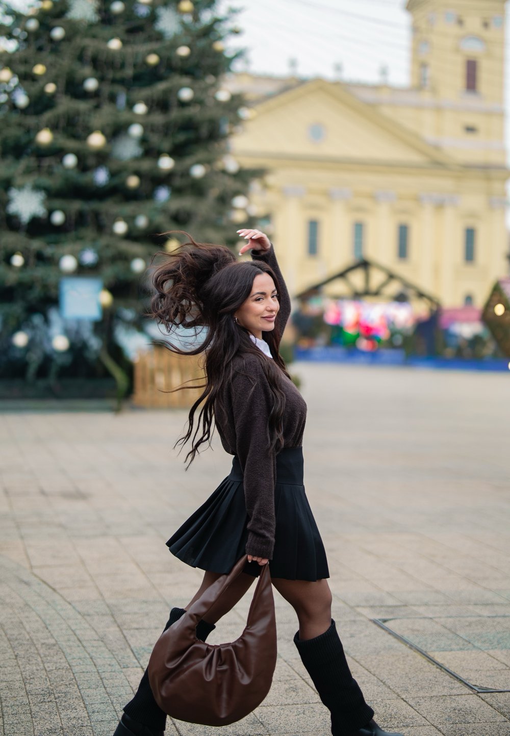 Sac en cuir marron souple, pull foncé, jupe plissée noire, chaussettes hautes noires, cheveux longs et ondulés, sapin de Noël enneigé en arrière-plan.