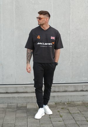 Hombre con tatuajes que lleva gafas de sol, camiseta negra del equipo McLaren de Fórmula 1, pantalones cargo negros y zapatillas blancas, de pie frente a una pared gris.