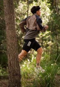 Mees jookseb metsaradadel, kandes tuulekindlat jope, musti triibulisi särki ja kõrvaklappe, ümbritsetuna rohelisest lehestikust ja kõrgest rohust.