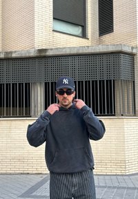 Σκούρο γκρι φούτερ με μπροστινή τσέπη, με απαλή υφή. Συνδυασμένο με παντελόνια με μαύρες και λευκές ρίγες. Μαύρο καπέλο και γυαλιά ηλίου.