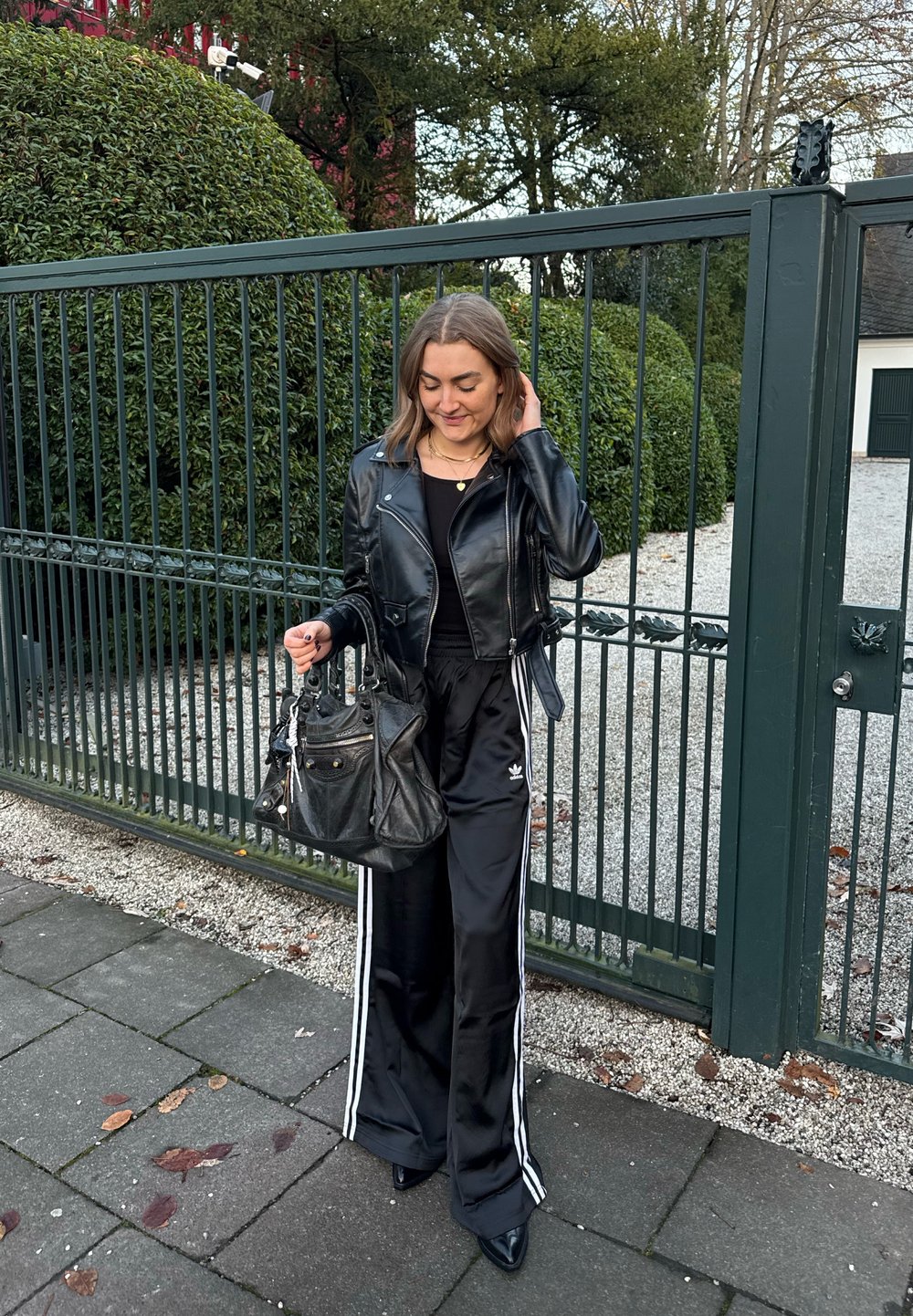 Veste en cuir noire, haut noir ajusté, pantalon en satin noir avec des rayures blanches, et chaussures à talons hauts. Porte un grand sac à main noir.