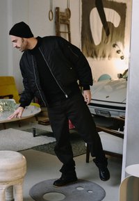 Chaqueta bomber negra sobre una camisa negra, combinada con pantalones oscuros y zapatos negros. De pie junto a una mesa redonda de color claro y muebles.