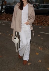 Beige ullfrakk med brede slag, hvit skjorte og hvite bukser. Brune støvler og en liten hvit skulderveske. Løv på bakken.