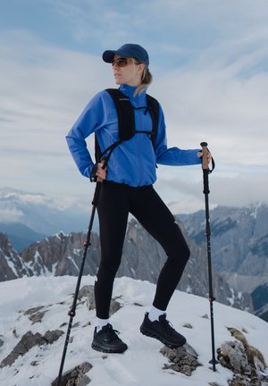 Giacca blu, leggings neri e scarpe nere con dettagli bianchi. Vengono utilizzati bastoncini da trekking e uno zaino. Paesaggio montuoso innevato e roccioso sullo sfondo.