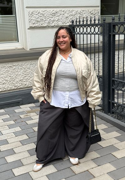 Persona sonriente con cabello largo trenzado que lleva una chaqueta beige, tops en capas, pantalones anchos de rayas finas, zapatos blancos y sostiene un bolso negro con cadena.