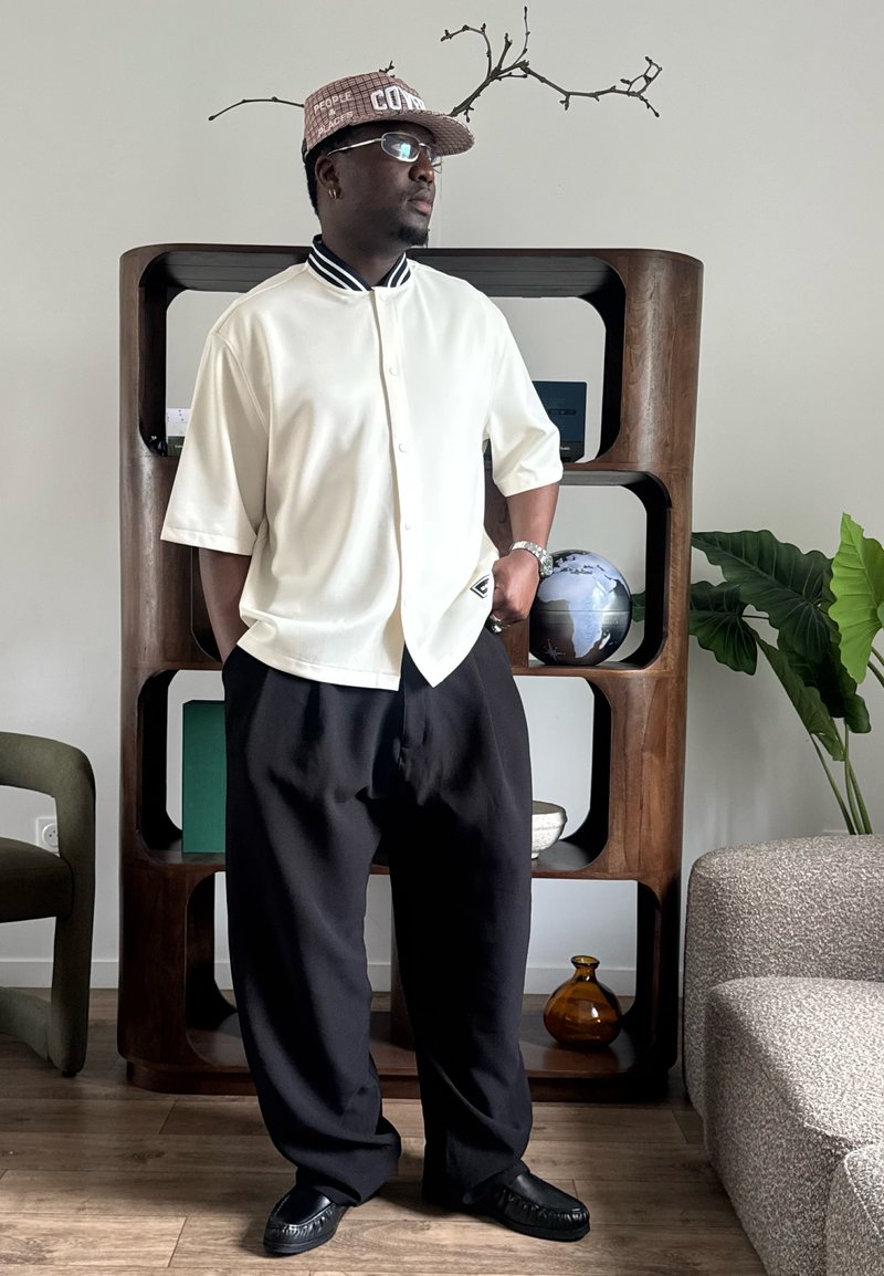 Homme portant une chemise blanche à manches courtes, pantalon noir et casquette à motif, debout dans une pièce moderne avec une étagère en bois, un globe et des plantes.