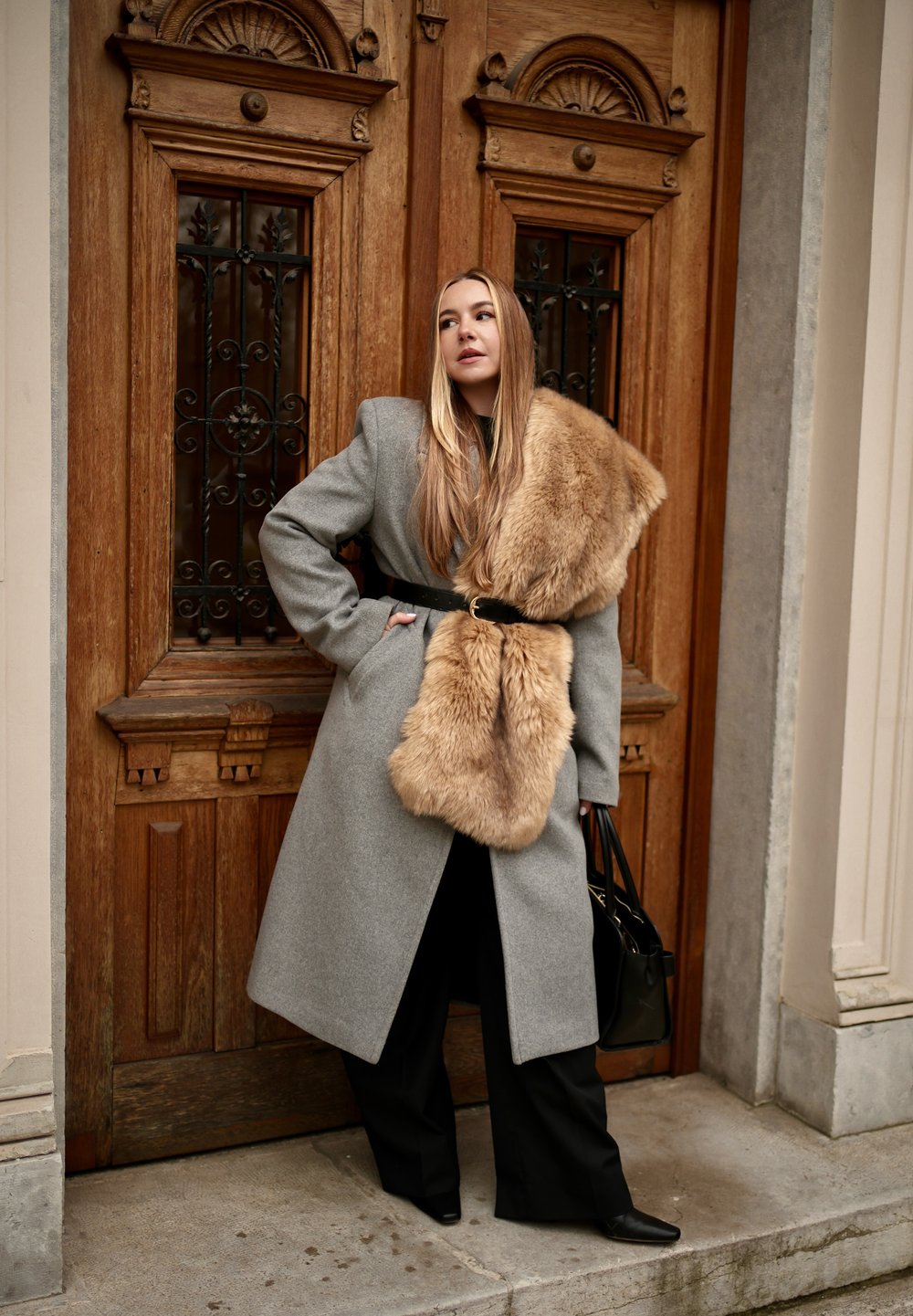 Femme portant un manteau gris ceinturé avec un grand col en fourrure camel, un pantalon noir et des chaussures noires, se tenant devant des portes doubles en bois ornées.