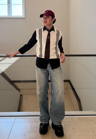 Chaleco de punto con rayas negras sobre crema, combinado con una camiseta negra, jeans anchos de lavado claro y zapatos negros. Una gorra burdeos completa el look.