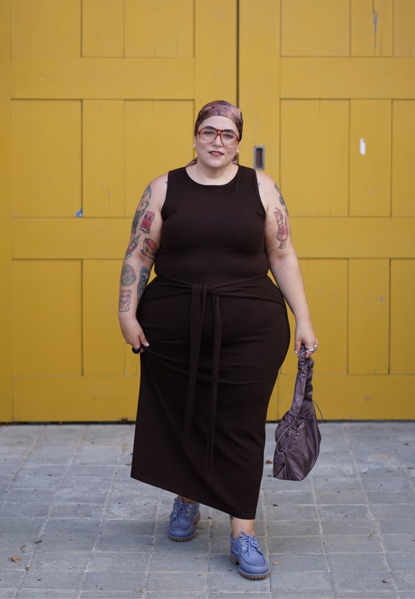 Personne grande taille avec des tatouages porte une robe noire sans manches, un foulard, des lunettes et des chaussures bleues, marchant sur le trottoir en tenant un petit sac.