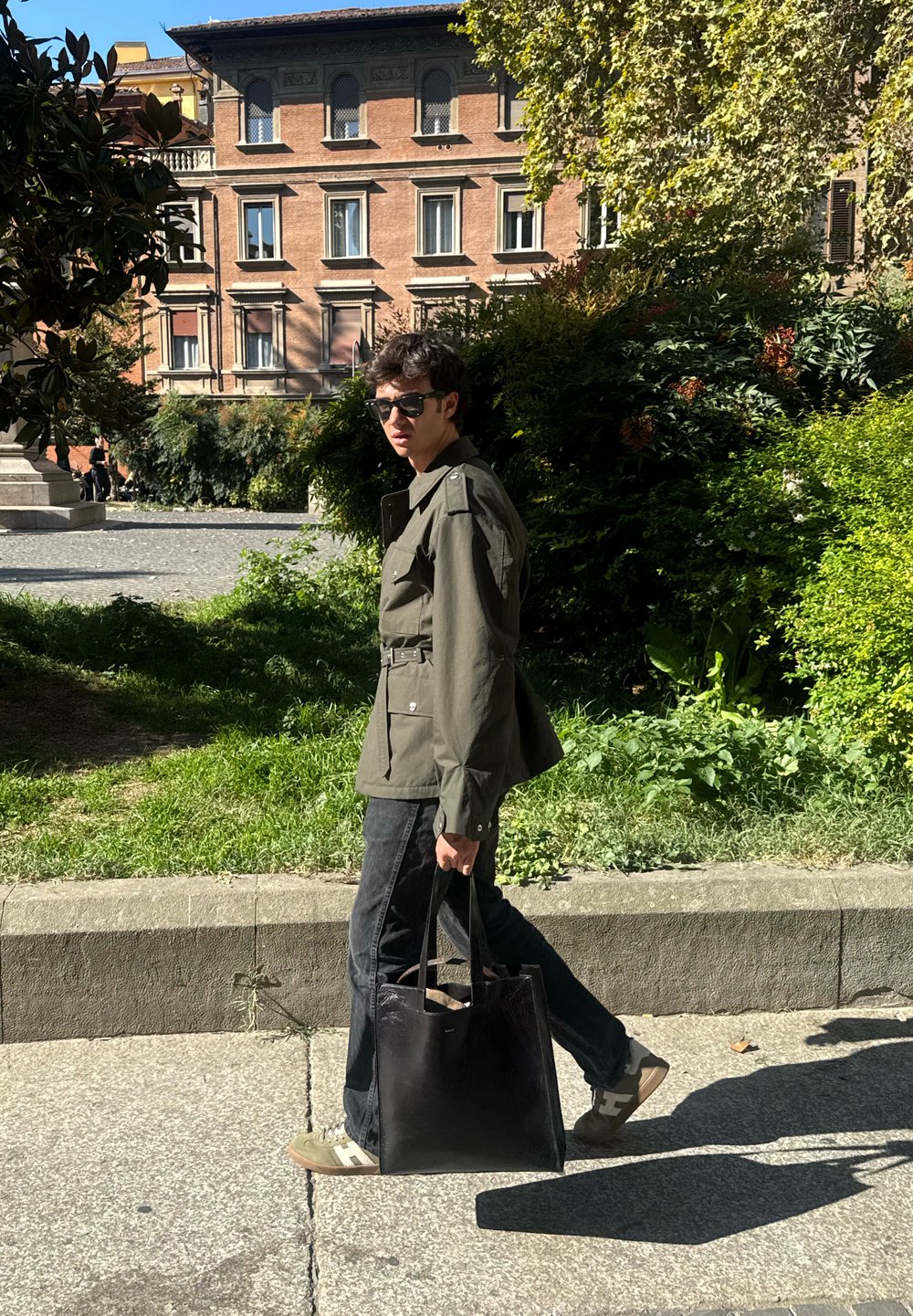 Olive green jacket with a belt, black jeans, black tote bag, and trainers. Natural light in a green outdoor setting with a building in the background.