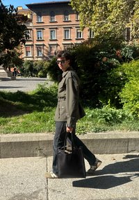 Olive green jacket with a belt, black jeans, black tote bag, and trainers. Natural light in a green outdoor setting with a building in the background.