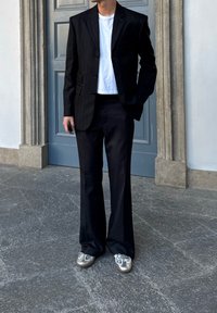 Čierne sako s jemnými pásikmi cez biele tričko, v kombinácii so zladenými čiernymi nohavicami a kovovo striebornými topánkami. Stojí na kamennom povrchu.