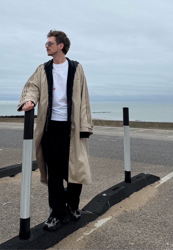Hombre con abrigo largo beige, pantalones negros y gafas de sol, apoyado en un poste blanco y negro cerca de un aparcamiento junto al mar bajo un cielo nublado.