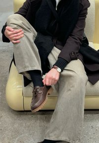 Scarpe da barca in pelle marrone con cuciture bianche, pantaloni di cady beige e una sciarpa nera a trama, seduto su un divano giallo pallido.