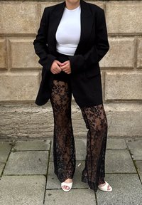 Blazer negro de gran tamaño, top blanco ajustado, pantalones de encaje negro transparente con patrón floral y tacones blancos abiertos en una superficie de piedra.