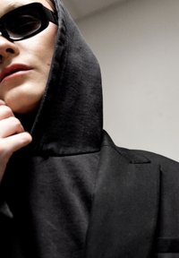 Sudadera con capucha negra de cuello alto y blazer negro. La persona lleva gafas de sol oscuras, con una textura textil suave y detalles mínimos.