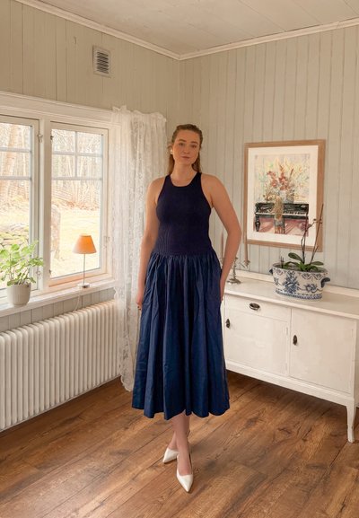 Mujer con top sin mangas azul marino y falda amplia de pie sobre suelo de madera cerca de una ventana con plantas en maceta y aparador con cuadro enmarcado.