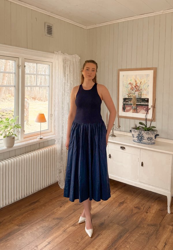 Donna con top blu navy senza maniche e gonna ampia in piedi su pavimento in legno vicino a finestra con piante in vaso e credenza con quadro incorniciato.