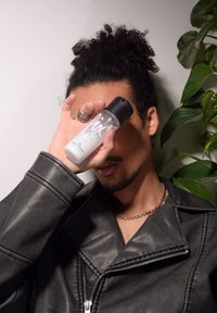 Bouteille vaporisateur transparente avec un capuchon noir, affichant du texte blanc. Une personne assise couvre partiellement son visage, portant une veste en cuir noire et un collier en chaîne.