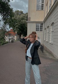 Μαύρο σακάκι πάνω από ένα ανοιχτό μπλε πουκάμισο, ανοιχτό γκρι παντελόνι και καφέ τσάντα. Το σκηνικό περιλαμβάνει λιθόστρωτο διάδρομο και μπεζ κτίρια.