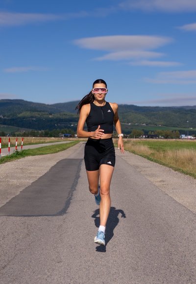 Conjunto deportivo negro con cremallera, con el logo de Adidas; gafas con montura rosa; zapatillas de running azules; corriendo por un camino pavimentado.