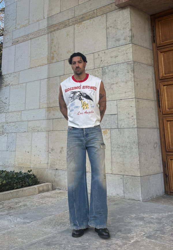 Homme aux cheveux foncés et tatouages portant un débardeur des Rolling Stones, un jean délavé à jambes larges et des chaussures noires, debout près d'un mur en pierre.