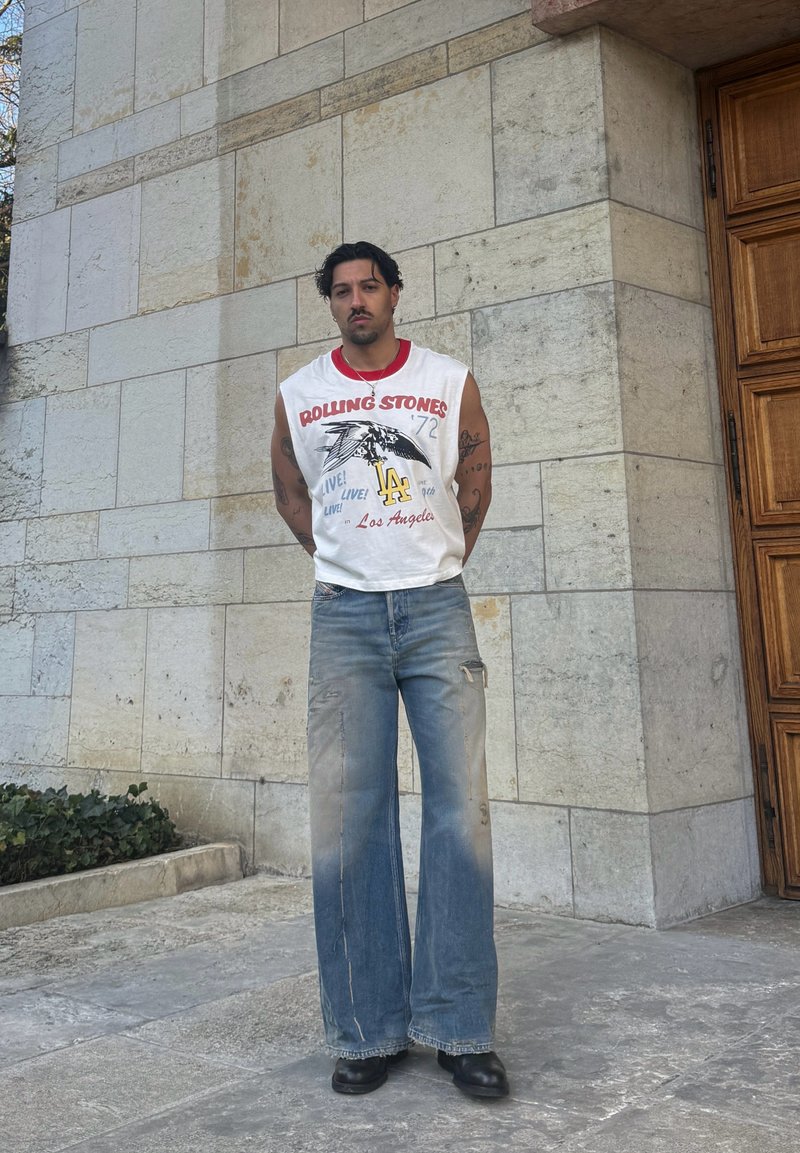 Άνδρας με σκούρα μαλλιά και τατουάζ, φορώντας αμάνικο μπλουζάκι των Rolling Stones, φαρδιά ξεθωριασμένα τζιν και μαύρα παπούτσια, στέκεται δίπλα σε πετρόχτιστο τοίχο.