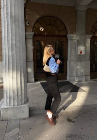 Camicia azzurra chiara, gilet nero, pantaloni neri, scarpe marroni e calzini bianchi. Piccola borsa beige. In piedi vicino a una colonna di pietra e porte decorative.