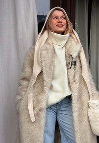 Femme portant un manteau en fausse fourrure beige clair avec une capuche, un pull à col roulé blanc et un jean bleu clair, debout près d'une fenêtre avec des rideaux.