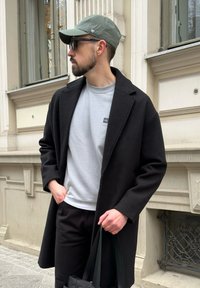 Melns vilnas mētelis pār pelēku džemperi, melnas bikses un melna soma rokā. Valkā zaļu cepuri un saulesbrilles. Pilsētas fonā.