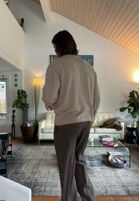 Person med brunt hår iført beige genser og mørke bukser som går bort fra stuen med hvit sofa, glasskaffebord og dekorative planter.