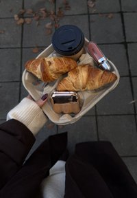 Brett med to gyllenbrune croissanter, en svart kaffekopp med lokk, et rosafarget etui og et rosa leppeprodukt på grått brostein.