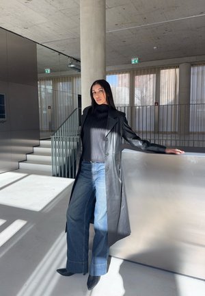 Mujer posando en interiores, apoyada en una superficie metálica, viste un abrigo largo de cuero negro, top negro, jeans de pierna ancha y botas negras puntiagudas.