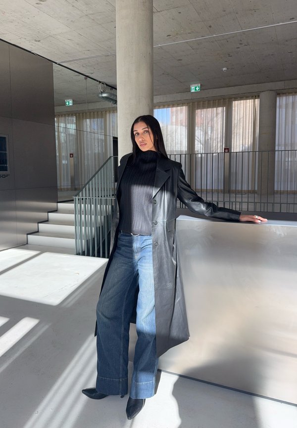 Donna in posa all'interno, appoggiata su una superficie metallica, indossa un lungo cappotto di pelle nero, una maglietta nera, jeans a gamba larga e stivali neri con punta appuntita.