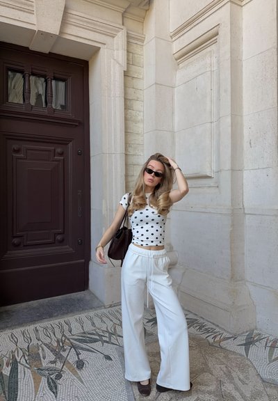 Joven mujer con cabello largo, lleva gafas de sol negras, una blusa blanca con lunares, pantalones blancos anchos y sostiene una bolsa negra junto a un edificio de piedra.
