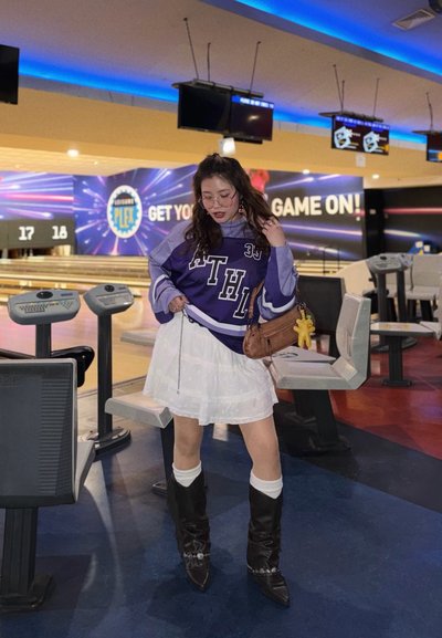 Joven mujer con camiseta deportiva morada y falda blanca posa en una bolera con pistas, monitores y un cartel que dice «¡A jugar!» en el fondo.