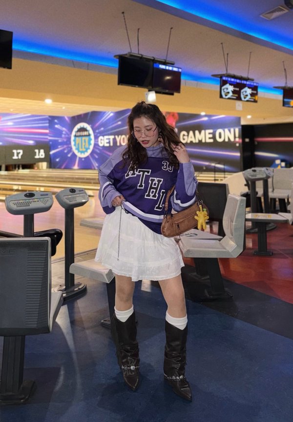 Giovane donna con maglietta sportiva viola e gonna bianca posa in una sala da bowling con piste, monitor e insegna "Game On!" sullo sfondo.