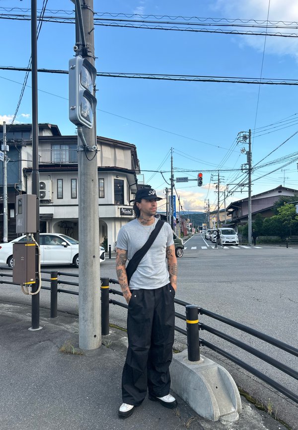 Hombre con tatuajes que lleva una camisa gris y pantalones negros, de pie en la esquina de la calle cerca de un semáforo que muestra la señal roja.